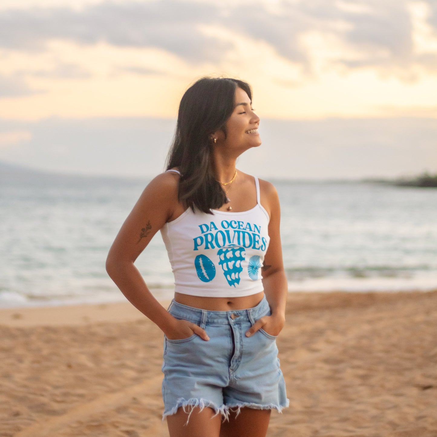 Woman standing on a beach with a sunset in the background, wearing a white tank top with text and denim shorts.