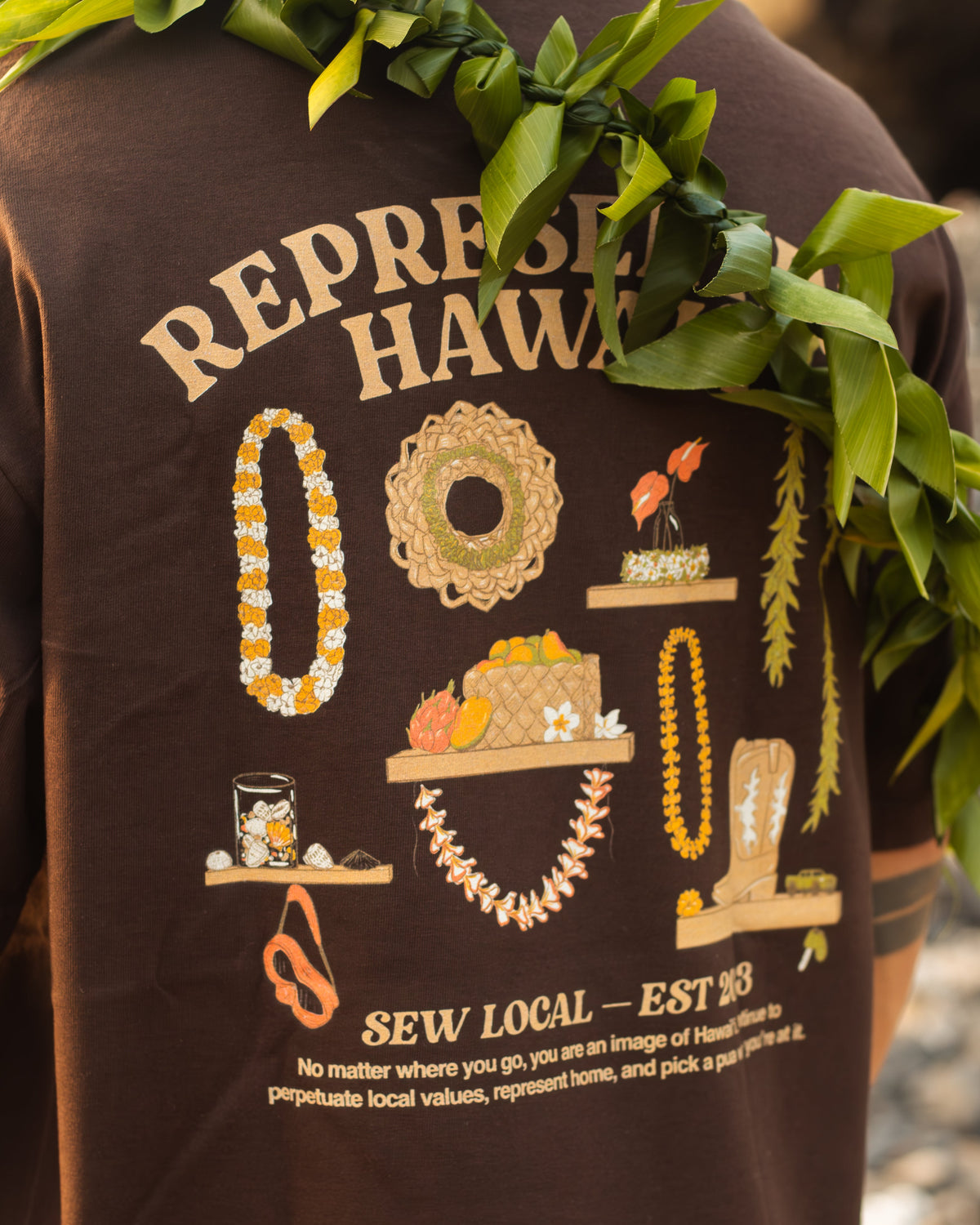 A close-up of a person wearing a dark brown graphic T-shirt outdoors, with green leaves draped over the shoulders. The back of the shirt features cream-colored text reading 'REPRESENT HAWAII' and 'SEW LOCAL - EST.