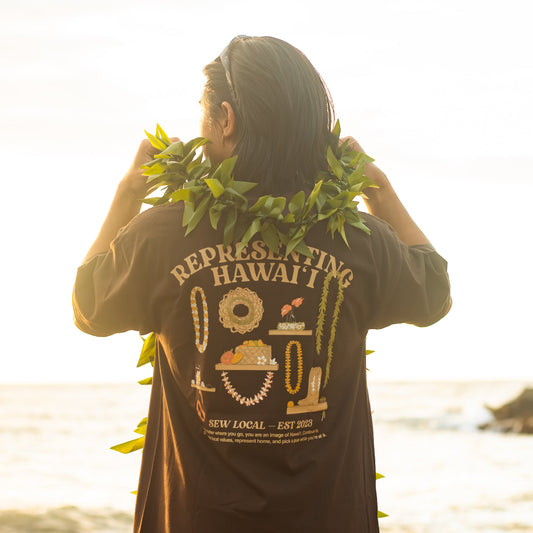 A person standing on a beach at sunset, viewed from behind, wearing a dark brown graphic T-shirt with 'REPRESENTING HAWAII' and Hawaiian-themed illustrations on the back, along with a green leaf lei.