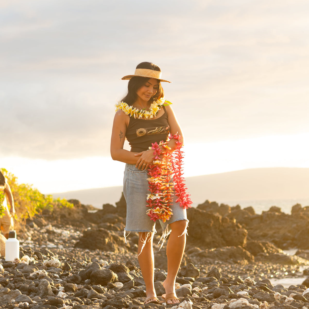 A woman stands on a rocky beach at sunset wearing a straw hat, a black graphic top, a light denim skirt, and holding bright orange and yellow flower leis. Another person is partially visible in the background.