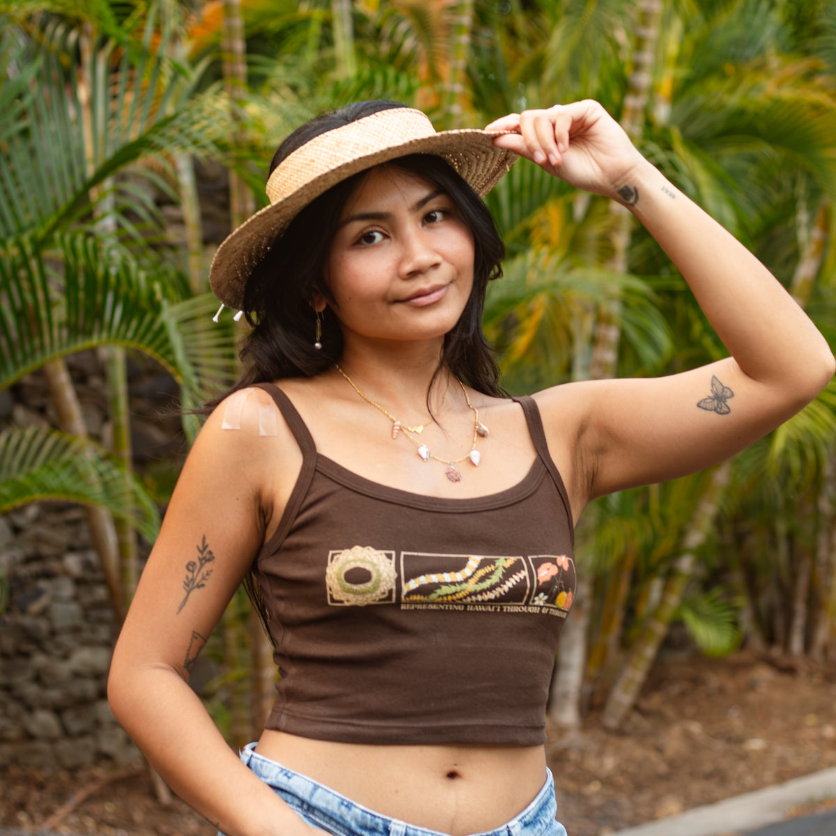 A woman wearing a brown cropped tank top with a graphic print, light blue jeans, and a straw hat, posing outdoors in front of a tropical background of palm trees.