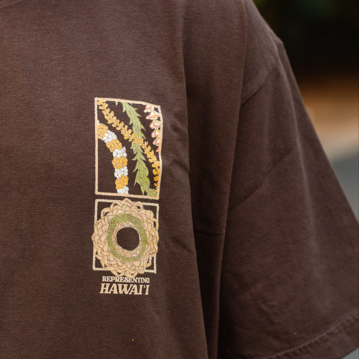 A close-up of a person wearing a dark brown short-sleeve t-shirt with a vertical graphic print on the left chest. The graphic features a split design: the top half shows green and yellow tropical leaves, and the bottom half shows a circular floral wreath with the word 'HAWAII' printed below.