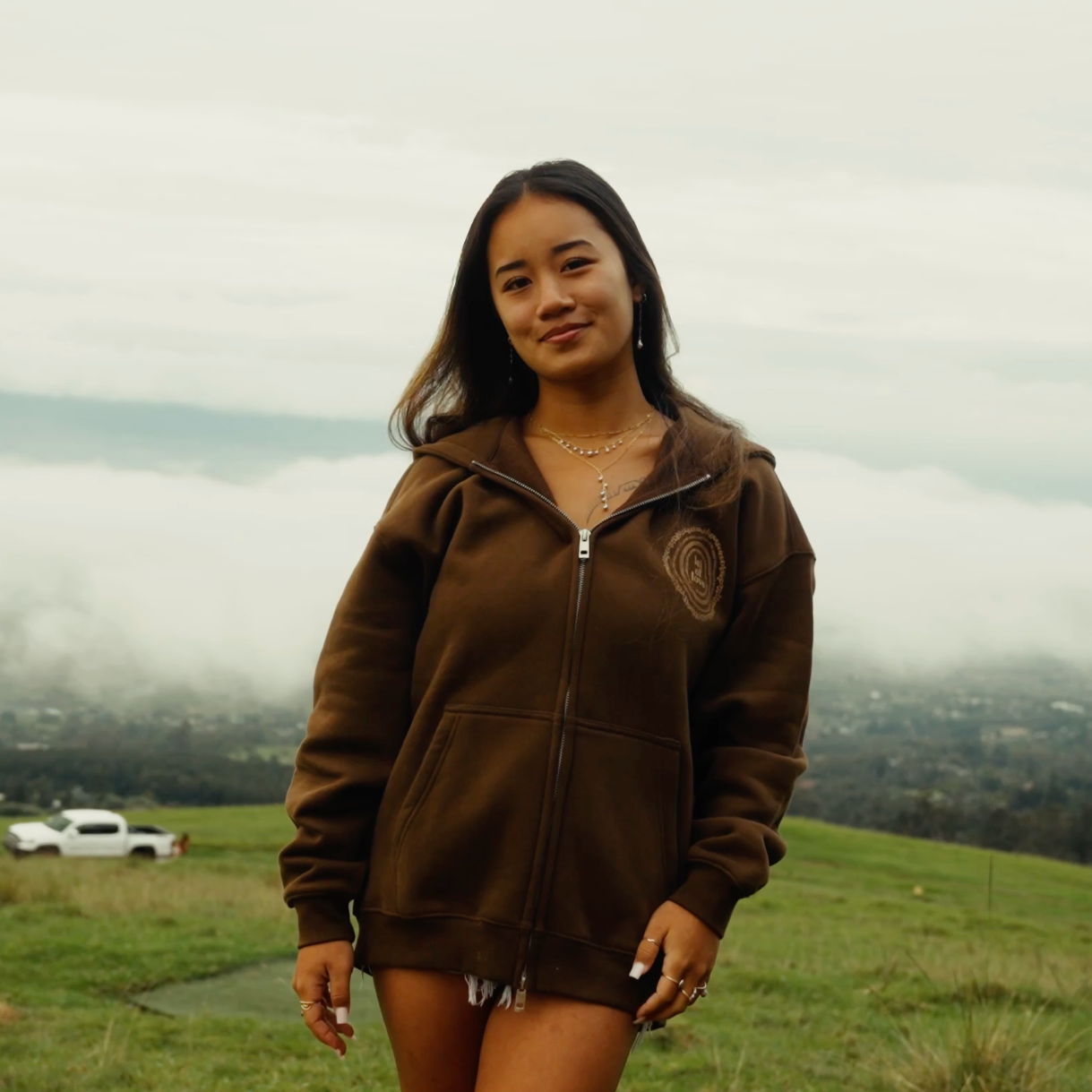 Woman wearing a brown hoodie and shorts standing in a grassy field with a cloudy sky.