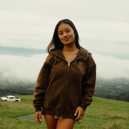 Woman wearing a brown hoodie and shorts standing in a grassy field with a cloudy sky.