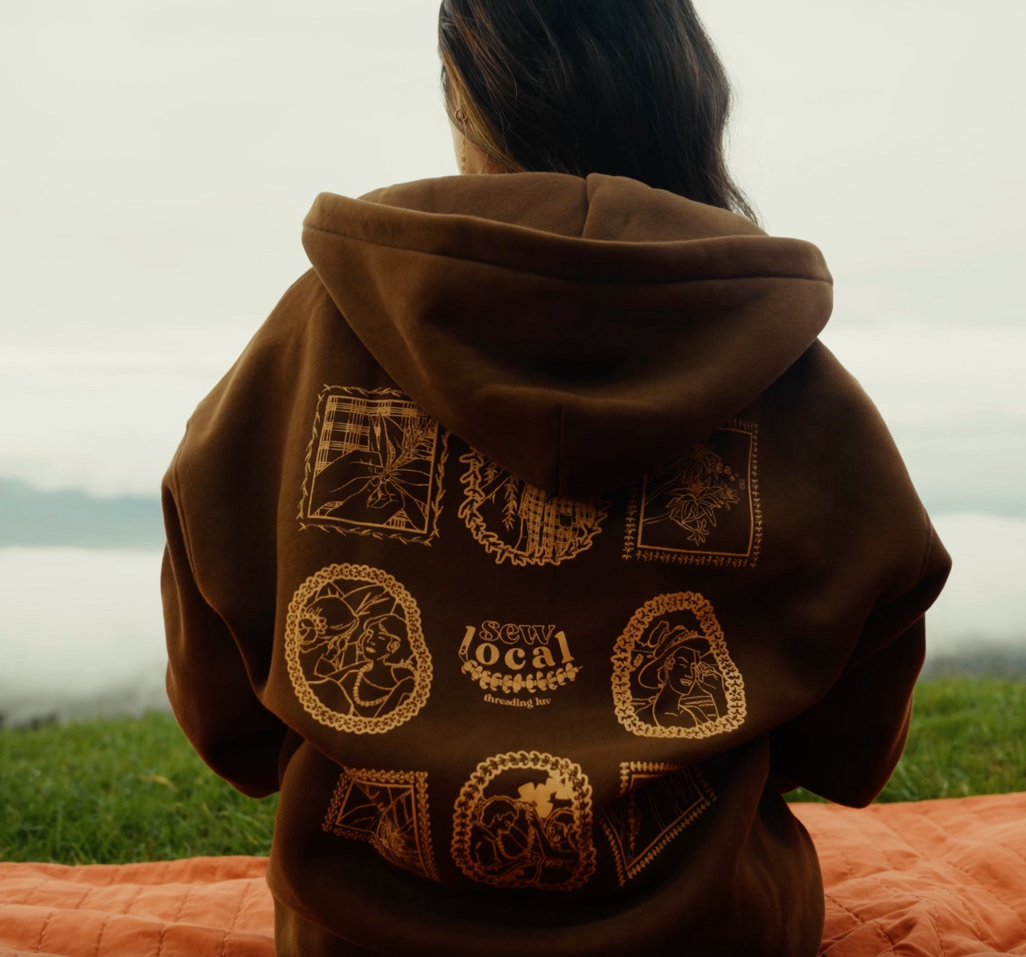 Person wearing a brown hoodie with lei of love designs, sitting on blanket