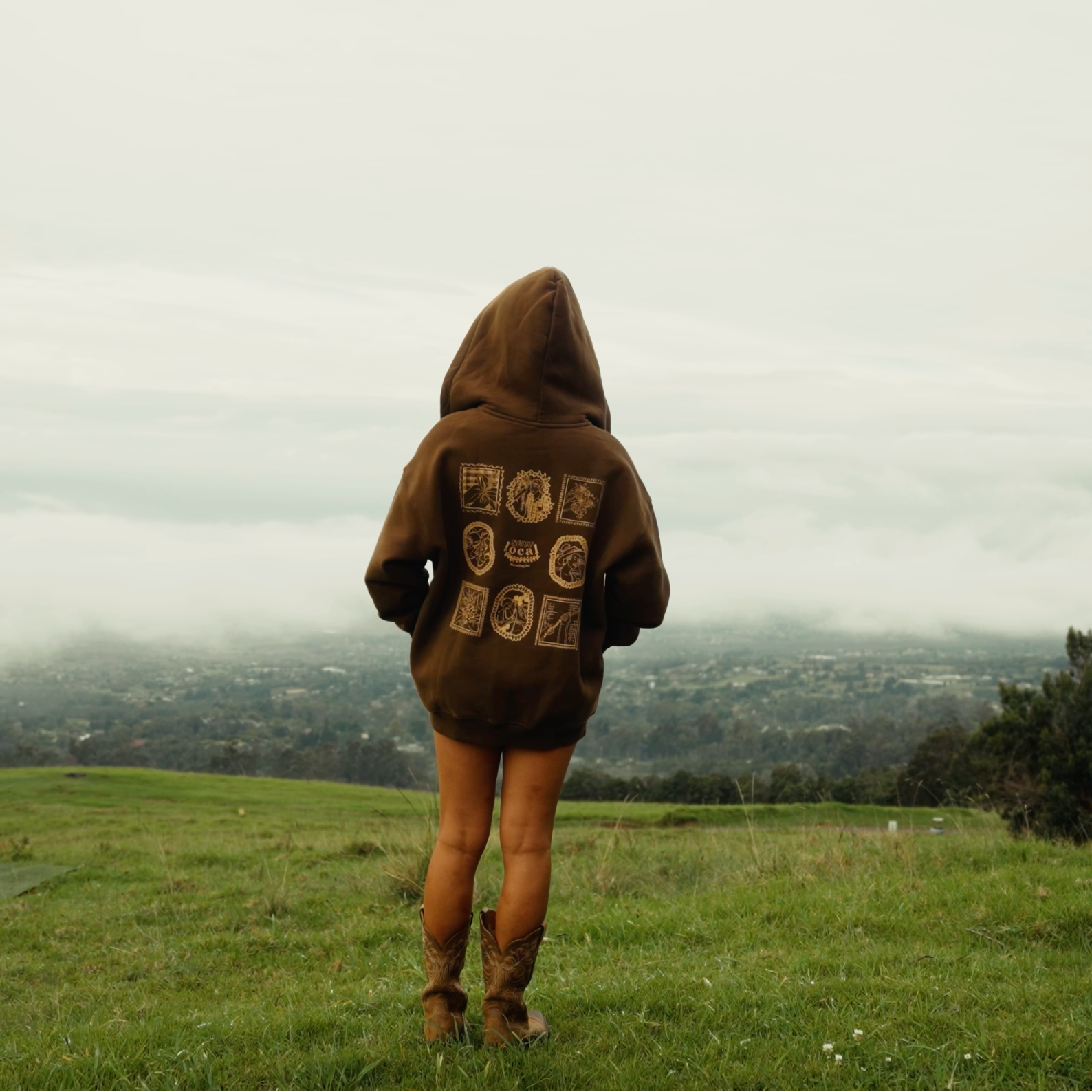 Person wearing a brown hoodie with text, standing in a grassy field with a cloudy sky.